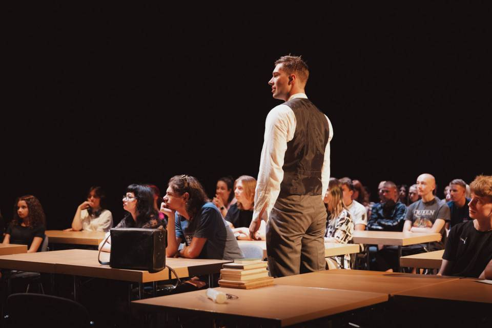 Ein Mann in Hemd und Weste steht in einem abgedunkelten Raum zwischen Reihen von Tischen, an denen viele junge Menschen sitzen. Die Szene wirkt wie eine Unterrichts- oder Theatervorstellung, mit Büchern und einer Tasche auf den Tischen im Vordergrund.