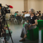 Eine Frau sitzt auf einem grünen Sitzwürfel und wird von einer Kamera mit Mikrofon interviewt. Im Hintergrund sitzen mehrere Menschen an Tischen in einem hellen Innenraum.