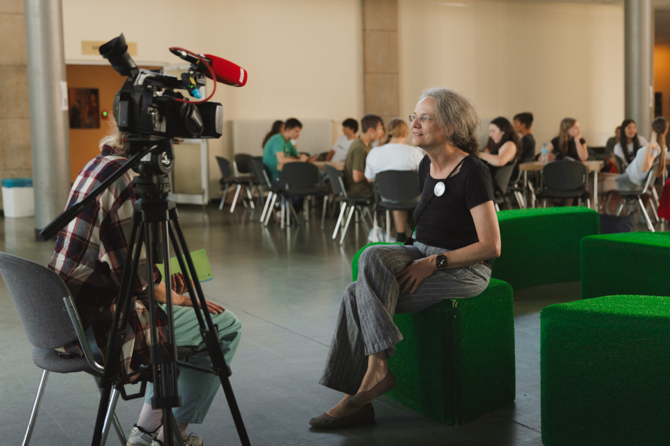 Eine Frau sitzt auf einem grünen Sitzwürfel und wird von einer Kamera mit Mikrofon interviewt. Im Hintergrund sitzen mehrere Menschen an Tischen in einem hellen Innenraum.