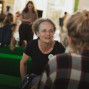 Eine Frau mit Brille sitzt auf einem grünen Sofa und spricht aufmerksam mit einer ihr gegenüber sitzenden Person. Im Hintergrund sind weitere Menschen in einem hellen, belebten Innenraum unscharf zu sehen.