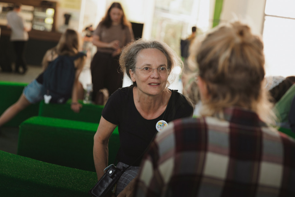 Eine Frau mit Brille sitzt auf einem grünen Sofa und spricht aufmerksam mit einer ihr gegenüber sitzenden Person. Im Hintergrund sind weitere Menschen in einem hellen, belebten Innenraum unscharf zu sehen.
