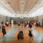 In einer großen Sporthalle tanzen viele Jugendliche und junge Erwachsene verteilt auf dem Spielfeld. Im Hintergrund sind ein Basketballkorb, Turngeräte und helle Fenster zu sehen.
