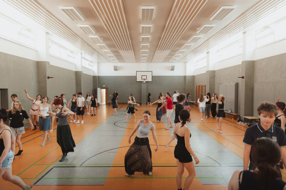 In einer großen Sporthalle tanzen viele Jugendliche und junge Erwachsene verteilt auf dem Spielfeld. Im Hintergrund sind ein Basketballkorb, Turngeräte und helle Fenster zu sehen.