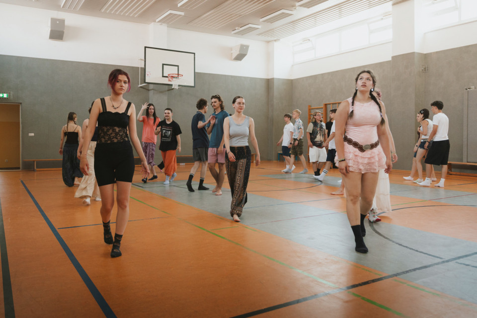 In einer hellen Sporthalle mit Basketballkorb laufen mehrere junge Menschen locker umher, teils barfuß oder in Socken. Im Vordergrund gehen drei Personen in auffälliger Freizeitkleidung auf die Kamera zu, während im Hintergrund weitere Gruppen miteinander interagieren.