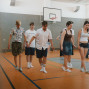 Eine Gruppe junger Menschen bewegt sich in einer Turnhalle über den orangefarbenen Sportboden, einige barfuß oder in Socken. Im Hintergrund sind ein Basketballkorb, graue Wände und weitere Personen zu sehen.