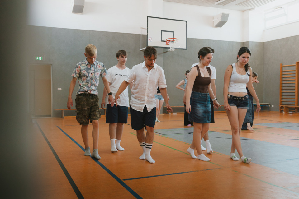 Eine Gruppe junger Menschen bewegt sich in einer Turnhalle über den orangefarbenen Sportboden, einige barfuß oder in Socken. Im Hintergrund sind ein Basketballkorb, graue Wände und weitere Personen zu sehen.