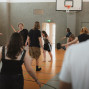 Mehrere Personen bewegen sich in einer Sporthalle, vermutlich bei einer Tanz- oder Bewegungsübung. Im Hintergrund sind ein Basketballkorb, Kletterstangen und eine geöffnete Tür zu sehen.