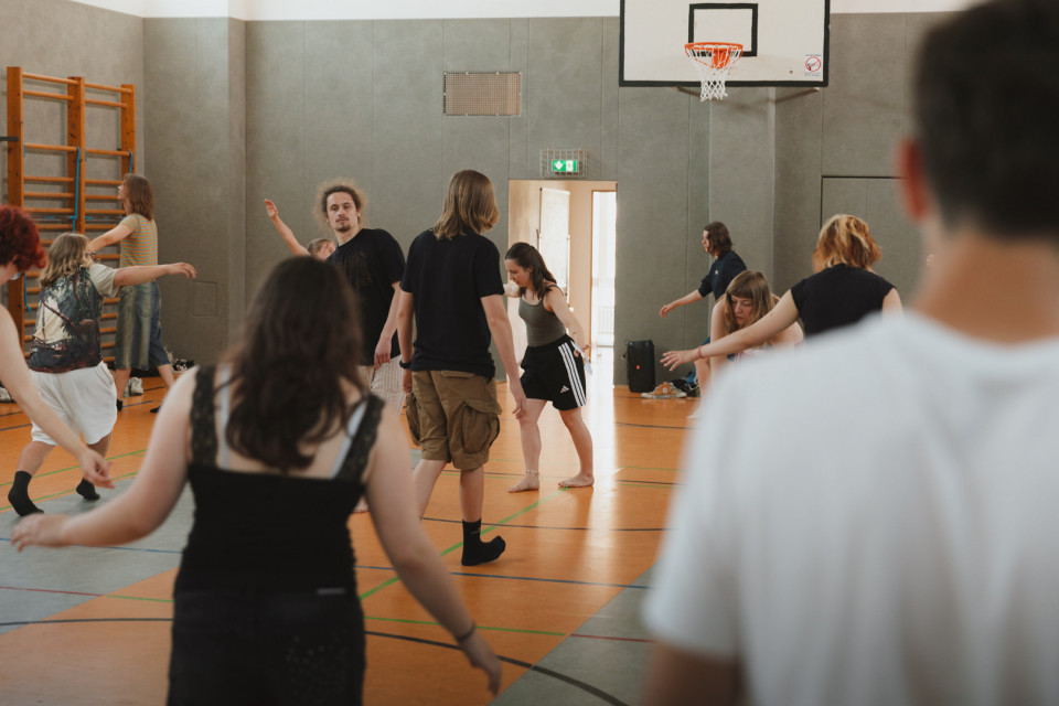 Mehrere Personen bewegen sich in einer Sporthalle, vermutlich bei einer Tanz- oder Bewegungsübung. Im Hintergrund sind ein Basketballkorb, Kletterstangen und eine geöffnete Tür zu sehen.