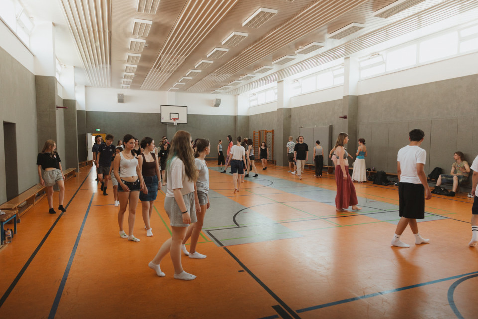 In einer hellen Sporthalle stehen und gehen viele junge Menschen in lockerer Kleidung und Socken auf dem orangefarbenen Hallenboden. An den Wänden sind Bänke, Sportgeräte und ein Basketballkorb zu sehen, während die Gruppe offenbar an einer gemeinsamen Übung oder Probe teilnimmt.