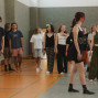 Eine Gruppe von Jugendlichen steht und geht locker verteilt in einer Sporthalle, einige barfuß oder in Socken. Im Hintergrund sind graue Wände, Bodenmarkierungen und ein Basketballkorb zu sehen.