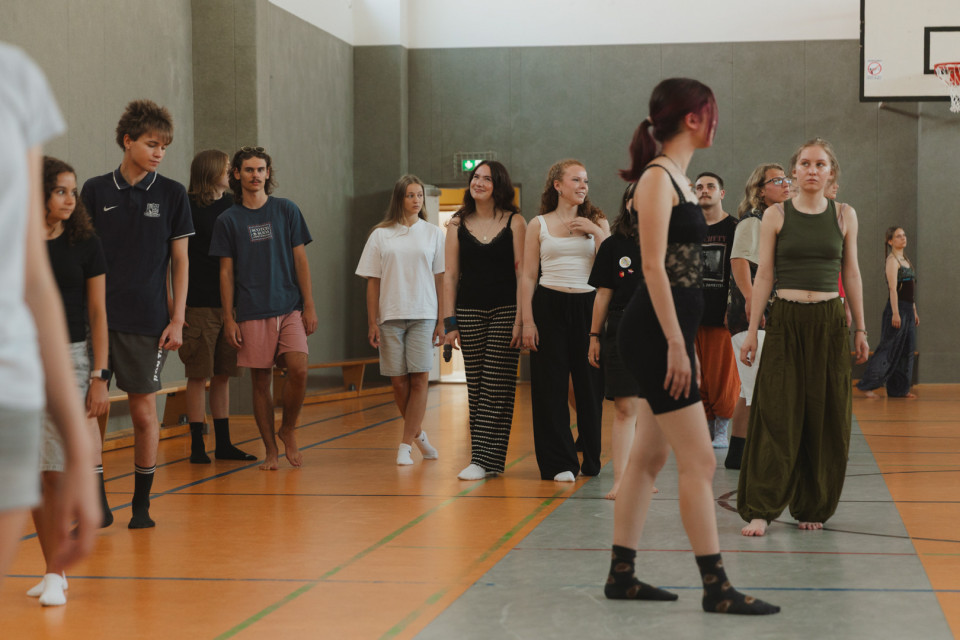 Eine Gruppe von Jugendlichen steht und geht locker verteilt in einer Sporthalle, einige barfuß oder in Socken. Im Hintergrund sind graue Wände, Bodenmarkierungen und ein Basketballkorb zu sehen.