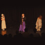 Auf einer dunklen Theaterbühne stehen drei kostümierte Kinder vor einem schwarzen Vorhang. Im Vordergrund sind die Silhouetten des Publikums zu sehen, während die Darsteller von warmem Bühnenlicht beleuchtet werden.