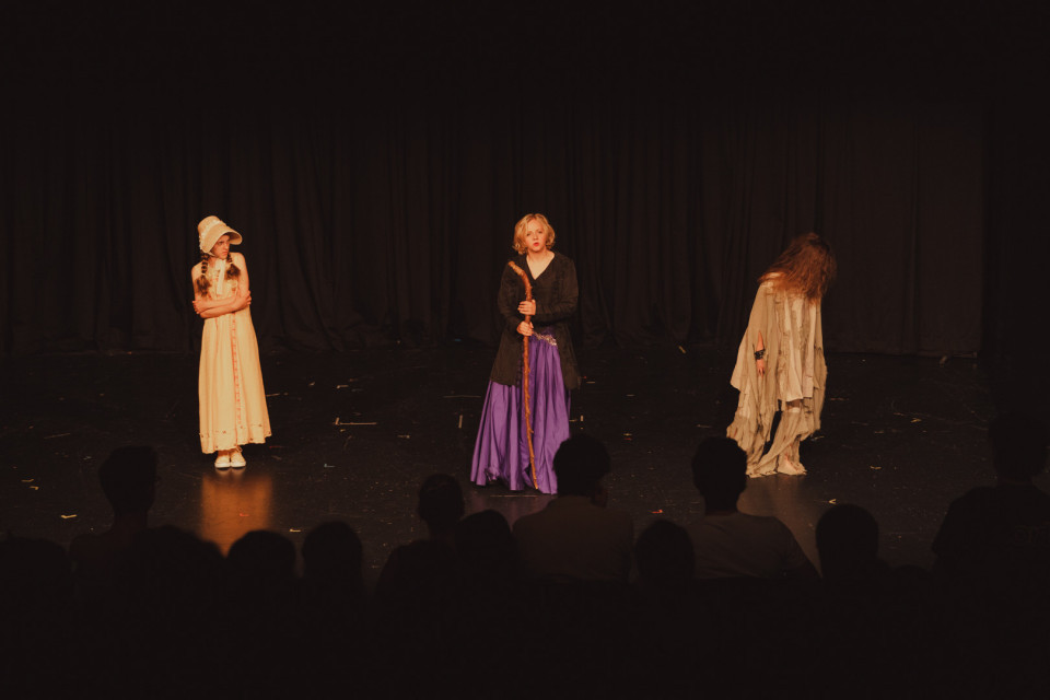 Auf einer dunklen Theaterbühne stehen drei kostümierte Kinder vor einem schwarzen Vorhang. Im Vordergrund sind die Silhouetten des Publikums zu sehen, während die Darsteller von warmem Bühnenlicht beleuchtet werden.