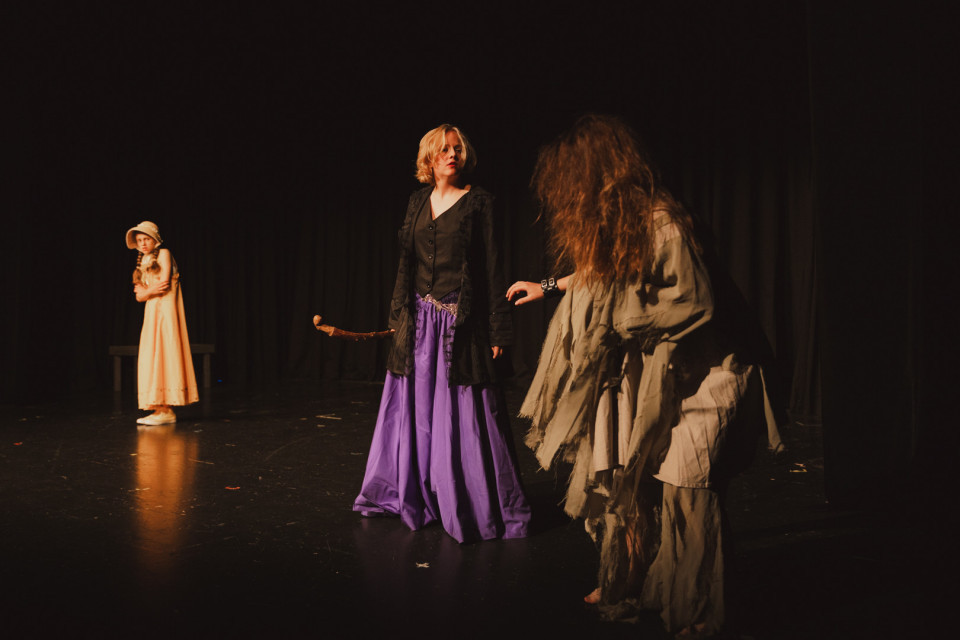 Auf einer dunklen Theaterbühne stehen drei kostümierte Personen im Scheinwerferlicht vor einem schwarzen Vorhang. In der Mitte trägt eine Figur einen schwarzen Mantel und einen violetten Rock, während rechts eine zerlumpte Gestalt mit langen Haaren und links eine Person in einem hellen Kleid zu sehen ist.