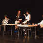 Auf einer dunklen Theaterbühne sitzen vier Personen an zwei Tischen und wirken wie in einer Szene oder Aufführung. Links hält ein Mann eine große Uhr-Requisite, während die anderen drei Personen auf Hockern sitzen und miteinander interagieren.