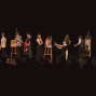 Auf einer dunklen Bühne stehen mehrere Menschen zwischen Staffeleien mit bunten Gemälden und wirken wie bei einer Kunst- oder Theateraufführung. Im Vordergrund sind die Silhouetten des Publikums zu sehen, während die Darstellerinnen und Darsteller miteinander interagieren.