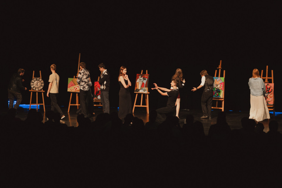 Auf einer dunklen Bühne stehen mehrere Menschen zwischen Staffeleien mit bunten Gemälden und wirken wie bei einer Kunst- oder Theateraufführung. Im Vordergrund sind die Silhouetten des Publikums zu sehen, während die Darstellerinnen und Darsteller miteinander interagieren.