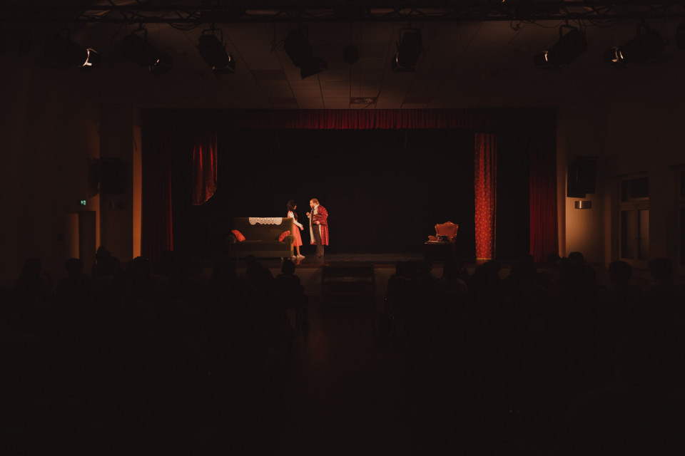 Auf einer dunkel beleuchteten Theaterbühne stehen zwei Schauspieler im Scheinwerferlicht vor einem roten Vorhang. Im Vordergrund sind die Zuschauer nur als dunkle Silhouetten zu erkennen, während auf der Bühne ein Sofa und ein Sessel als Kulisse stehen.