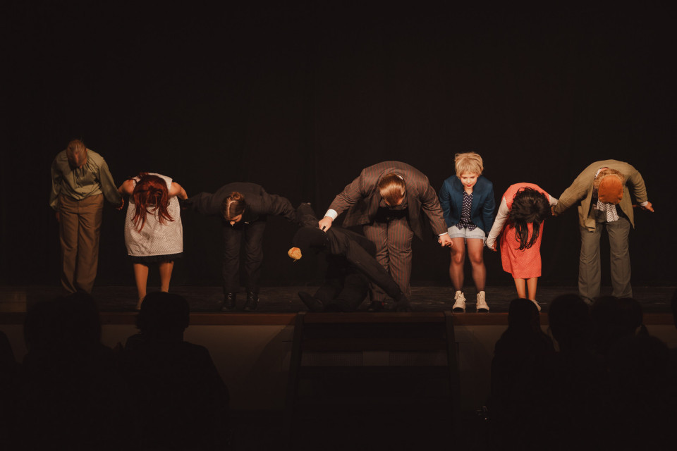 Eine Gruppe von Schauspielerinnen und Schauspielern verbeugt sich Hand in Hand auf einer dunkel beleuchteten Bühne. Im Vordergrund sind die Silhouetten des Publikums zu sehen, das der Aufführung zuschaut.
