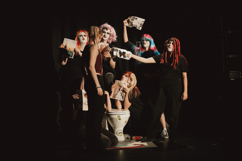 Auf einer dunklen Bühne posieren mehrere Personen mit auffälligem Make-up, bunten Perücken und schwarzen Outfits, während sie Zeitschriften und Blätter hochhalten. In der Mitte steht eine Toilette, um die sich die Gruppe theatralisch versammelt.