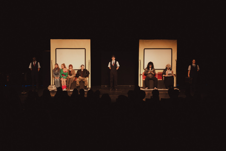 Auf einer dunklen Theaterbühne sind zwei beleuchtete, wie Zugabteile wirkende Sitzbereiche mit mehreren Darstellern zu sehen. Im Vordergrund erkennt man die Silhouetten des Publikums, während weitere Schauspieler in Uniformen seitlich und in der Mitte stehen.