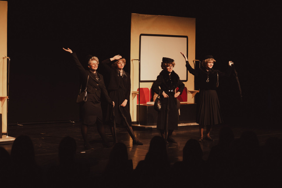 Auf einer Theaterbühne stehen vier schwarz gekleidete Darstellerinnen in eleganten Kostümen und posieren mit erhobenen Armen vor einem Bühnenbild, das wie ein Zugabteil wirkt. Im Vordergrund sind die dunklen Silhouetten des Publikums zu sehen, während warmes Licht die Szene beleuchtet.