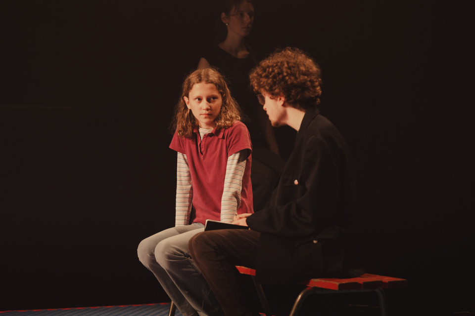 Auf einer dunkel beleuchteten Bühne sitzen ein Mädchen im roten Shirt und ein junger Mann einander gegenüber und schauen sich an. Im Hintergrund steht schemenhaft eine weitere Person, während warmes Licht die Szene dramatisch hervorhebt.
