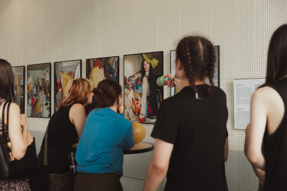 Eine Gruppe von Menschen betrachtet in einer Galerie mehrere gerahmte Fotografien an einer perforierten Wand. Eine Person zeigt auf eines der Bilder, während die anderen aufmerksam davor stehen.