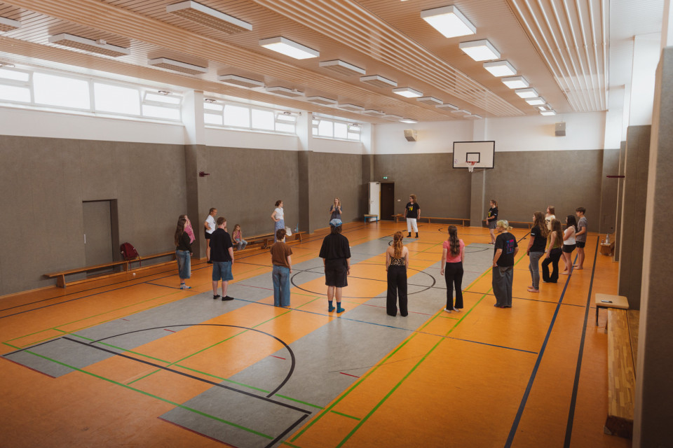 In einer großen Sporthalle stehen mehrere Jugendliche und Erwachsene in einem weiten Kreis auf dem Spielfeld. An der Wand hängt ein Basketballkorb, und die Halle ist hell beleuchtet mit hohen Fenstern und Holzbänken an den Seiten.