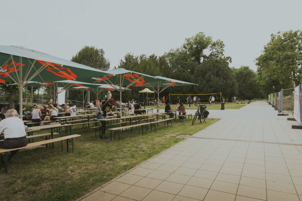 Auf einer Grünfläche sitzen Menschen an langen Bierbänken unter großen Sonnenschirmen. Im Hintergrund spielen einige Personen Volleyball, während rechts ein breiter gepflasterter Weg entlangführt.