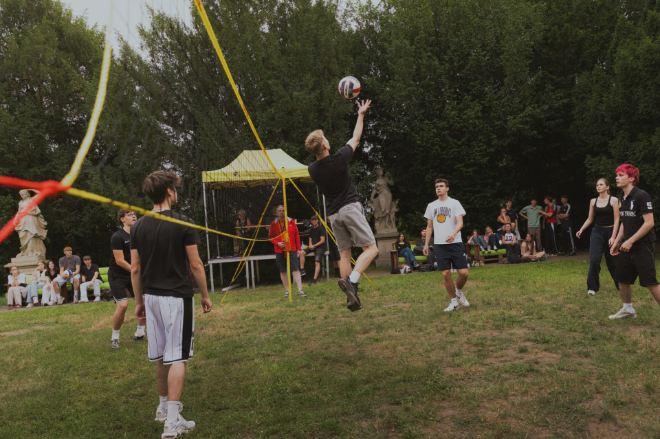 Eine Gruppe junger Menschen spielt im Park auf einer Wiese Volleyball, während ein Spieler am Netz hochspringt und den Ball schlägt. Im Hintergrund sitzen Zuschauer auf Bänken, daneben stehen Bäume, Statuen und ein gelbes Zelt.