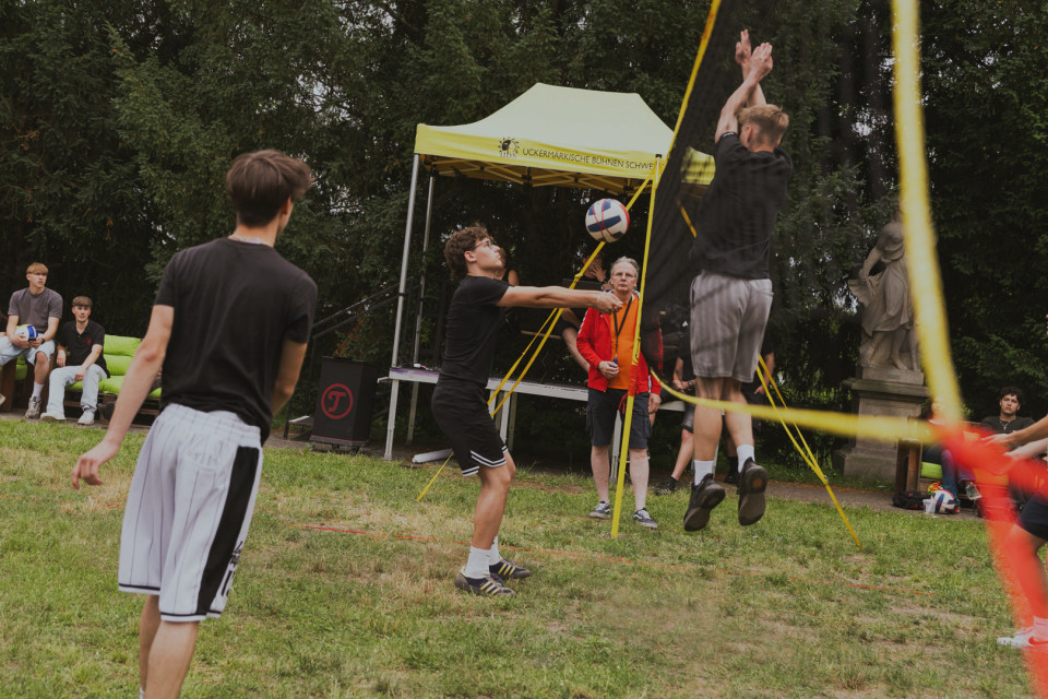 Mehrere junge Männer spielen draußen auf einer Wiese Volleyball, während ein Spieler am Netz hochspringt und den Ball blockt. Im Hintergrund stehen Zuschauer, ein gelbes Zelt und Bäume, wodurch die Szene wie ein kleines Freizeit- oder Turnierereignis wirkt.