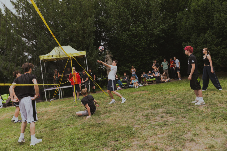 Eine Gruppe junger Leute spielt draußen auf einer Wiese Volleyball, während mehrere Zuschauer am Rand sitzen und zusehen. Im Hintergrund stehen Bäume sowie ein gelbes Zelt mit Netz, und der Ball fliegt gerade über das Spielfeld.