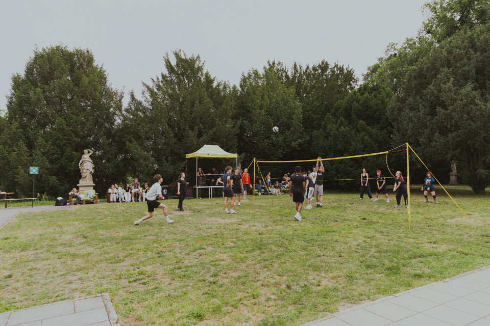 Mehrere Menschen spielen auf einer Wiese in einem Park Volleyball an einem gelben Netz. Im Hintergrund sitzen Zuschauer neben einem kleinen Pavillon, umgeben von Bäumen und Statuen.