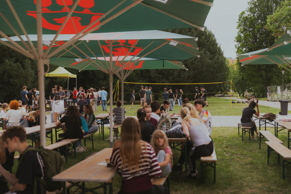 Viele Menschen sitzen draußen an Biertischen unter großen grünen Sonnenschirmen und unterhalten sich. Im Hintergrund spielen einige Personen auf einer Wiese Volleyball, während weitere Gruppen beisammenstehen.