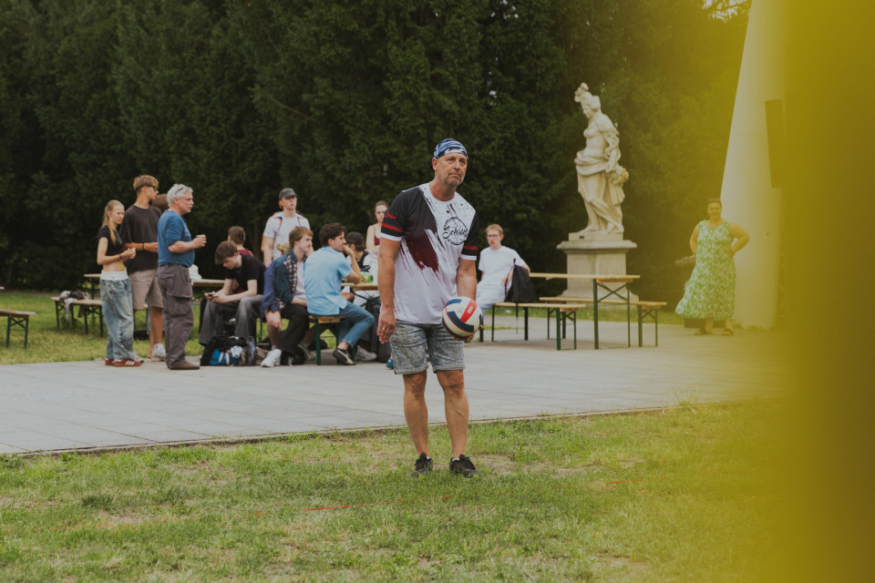 Ein Mann steht auf einer Wiese und hält einen Volleyball in der Hand, während im Hintergrund mehrere Personen an Biertischen sitzen oder stehen. Hinter ihnen sind ein Wald, eine Statue und ein gelblicher unscharfer Vordergrund zu sehen.