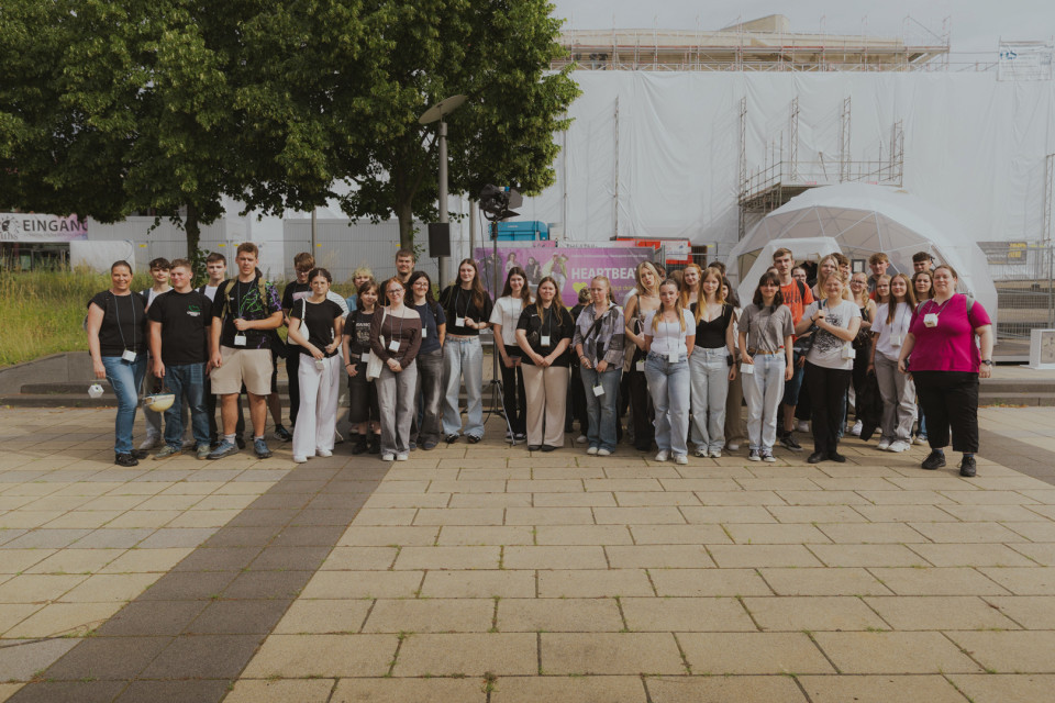 Eine große Gruppe junger Menschen steht mit Namensschildern auf einem gepflasterten Platz und posiert für ein Foto. Im Hintergrund sind Bäume, ein weiß verhülltes Gebäude mit Gerüsten sowie eine kuppelförmige Struktur zu sehen.