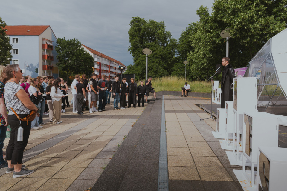 Eine Gruppe von Menschen steht auf einem gepflasterten Platz und hört einer Person zu, die auf einer kleinen Bühne an einem Mikrofon spricht. Im Hintergrund sind Wohnhäuser, Bäume und ein bewölkter Himmel zu sehen, während rechts weiße Ausstellungsobjekte und ein transparenter Pavillon stehen.