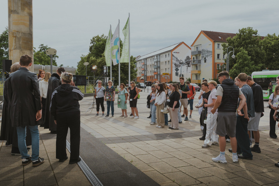 Eine Gruppe von Menschen versammelt sich auf einem städtischen Platz vor einem Gebäude, während links offenbar eine Ansprache oder Vorführung stattfindet. Im Hintergrund sind Fahnen, Bäume, Wohnhäuser und ein Bus zu sehen.