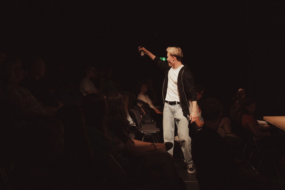 Ein junger Mann steht im Scheinwerferlicht zwischen den Zuschauerreihen eines dunklen Theaters und hält einen kleinen Gegenstand in die Höhe. Um ihn herum sitzen viele Menschen im Halbdunkel und beobachten die Szene.
