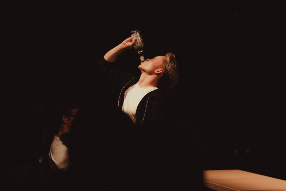 Ein Mann steht in einem dunklen Raum im Scheinwerferlicht und trinkt mit zurückgelegtem Kopf aus einer Flasche. Im Vordergrund sind dunkle Silhouetten von Zuschauern zu sehen, während rechts ein beleuchteter Tisch steht.