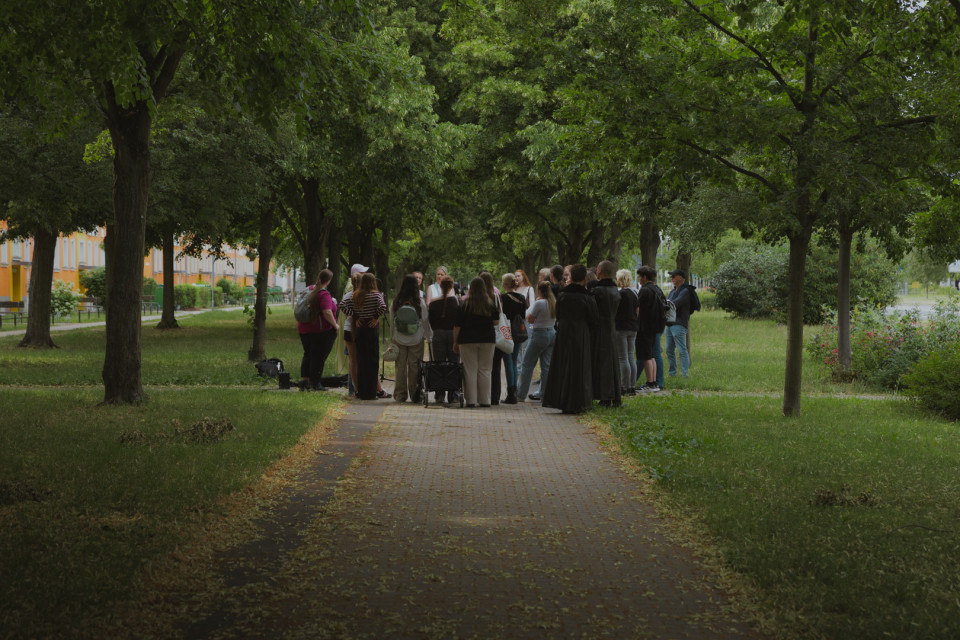 Eine Gruppe von Menschen steht auf einem gepflasterten Weg in einem grünen, von Bäumen gesäumten Park. Die Szene wirkt ruhig und schattig, während die Personen offenbar miteinander sprechen oder einer Führung zuhören.