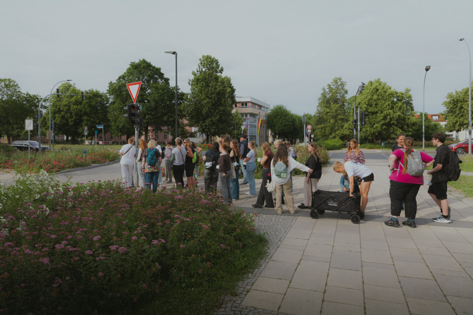 Eine größere Gruppe von Menschen wartet an einer Ampel an einer Straßenkreuzung. Im Vordergrund sind Blumenbeete zu sehen, während im Hintergrund Bäume, Verkehrsschilder und Gebäude stehen.