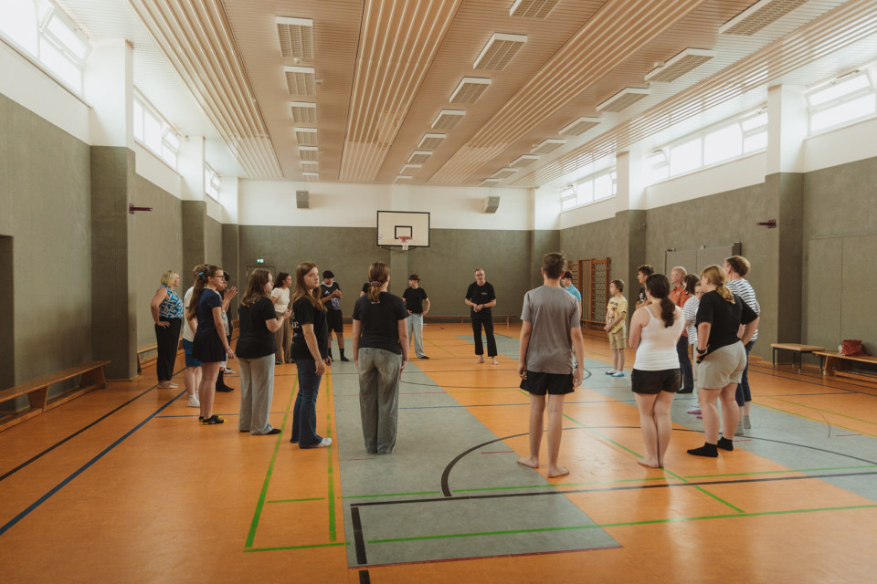 Eine Gruppe von Menschen steht in einer großen Sporthalle im Kreis und hört einer Person in der Mitte zu. Die Halle ist hell beleuchtet, hat hohe Fenster, einen Basketballkorb und markierte Spielfeldlinien auf dem Boden.