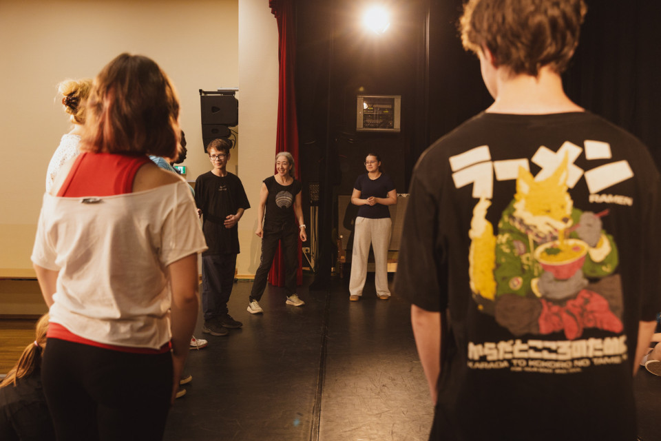 Eine Gruppe von Menschen steht auf einer Bühne oder in einem Proberaum und wirkt, als würde sie an einer Theater- oder Workshopübung teilnehmen. Im Vordergrund sind zwei Personen von hinten zu sehen, während in der Mitte mehrere Teilnehmende unter warmem Bühnenlicht stehen.