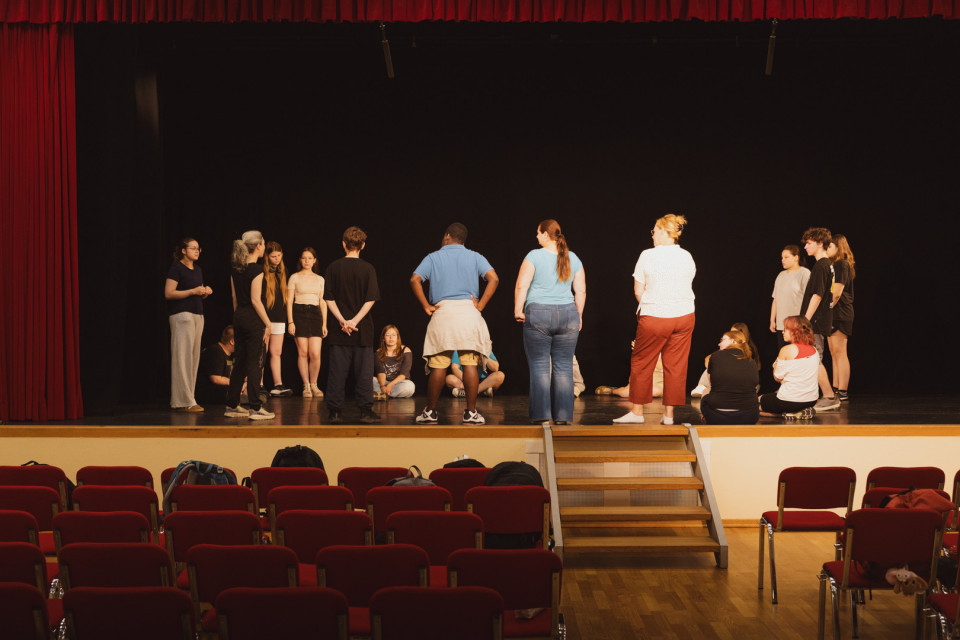 Eine Gruppe von Jugendlichen und Erwachsenen steht und sitzt auf einer Theaterbühne vor einem schwarzen Hintergrund und roten Vorhängen. Im Vordergrund sind leere rote Zuschauerstühle und eine Treppe zur Bühne zu sehen.