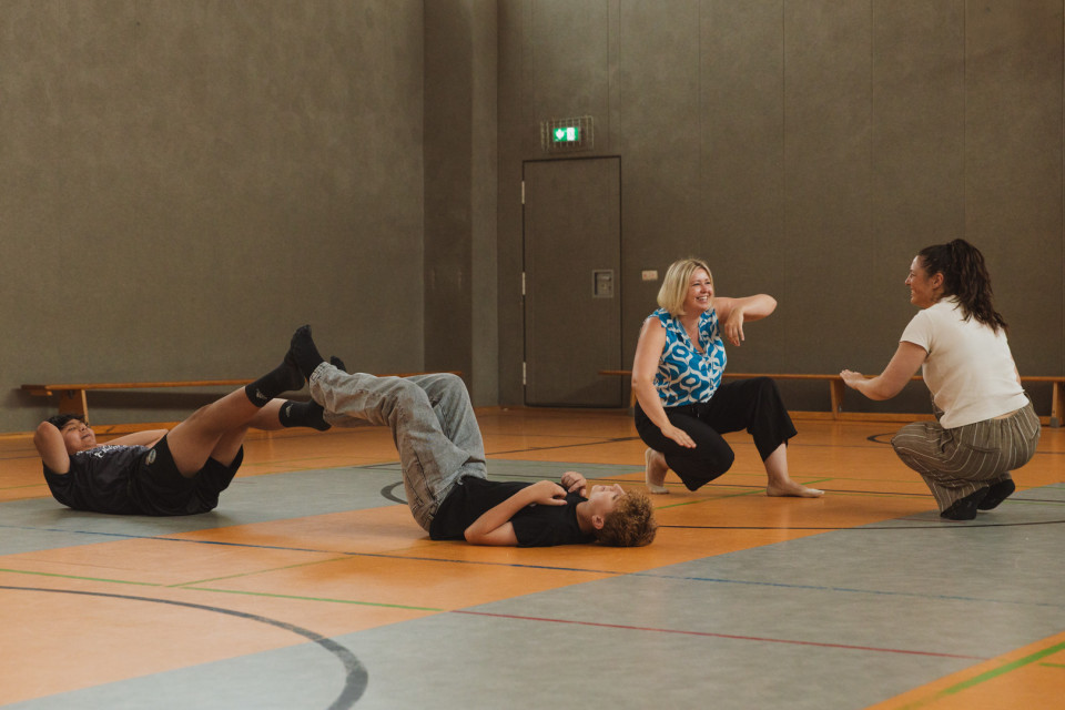 In einer Sporthalle liegen zwei Kinder spielerisch auf dem Boden und heben die Beine, während sie offenbar eine Übung machen. Daneben hocken zwei Frauen und unterhalten sich lachend, eine davon gestikuliert lebhaft.