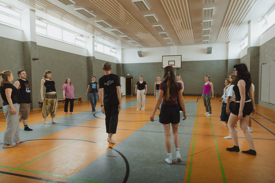 Eine Gruppe von Menschen steht in einer Sporthalle im Kreis, offenbar bei einer Tanz- oder Bewegungsübung. Die helle Halle hat markierte Spielfeldlinien, eine Basketballkorbwand und hohe Fenster.