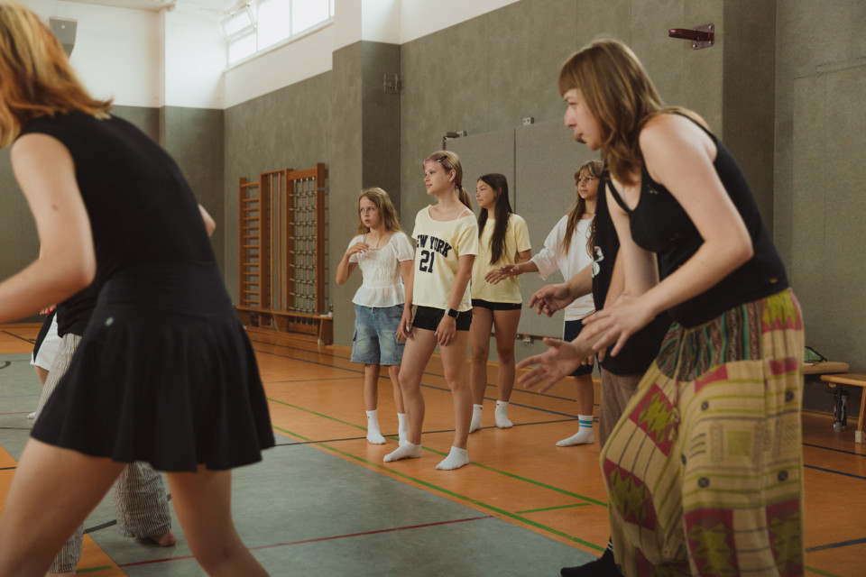 Mehrere Mädchen und junge Frauen stehen in einer Sporthalle und wirken, als würden sie an einer Bewegungs- oder Tanzübung teilnehmen. Im Vordergrund sind zwei Personen teilweise zu sehen, während die Gruppe im Hintergrund aufmerksam nach vorne schaut.