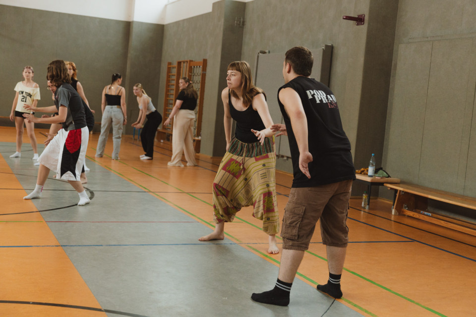 Mehrere Personen üben in einer Turnhalle gemeinsam Bewegungen oder Tanzschritte. Im Vordergrund stehen eine barfüßige Frau in einer gemusterten Hose und ein Mann im schwarzen ärmellosen Shirt einander gegenüber.